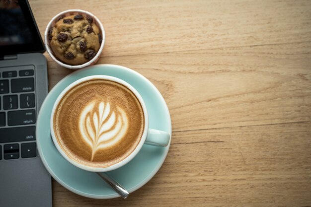 Primer plano de café con leche caliente con espuma de leche latte art en taza taza y pastel de taza de plátano casero con computadora portátil en escritorio de madera escritorio de oficina en cafetería en la cafetería, durante el concepto de trabajo empresarial