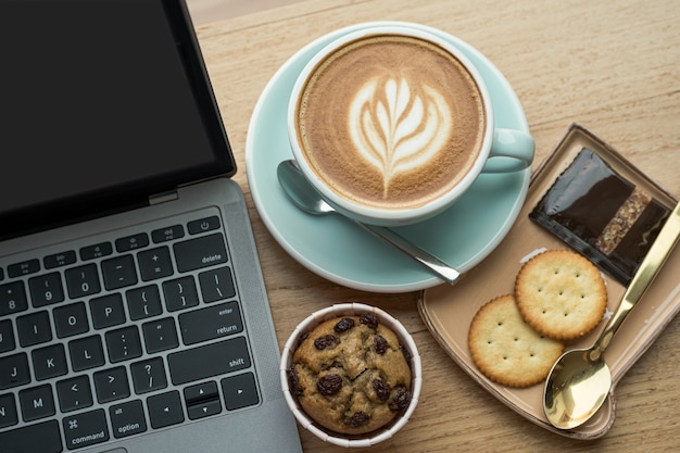 Primer plano de café con leche caliente con espuma de leche latte art en taza taza y pastel de taza de plátano casero con computadora portátil en escritorio de madera escritorio de oficina en cafetería en la cafetería, durante el concepto de trabajo empresarial