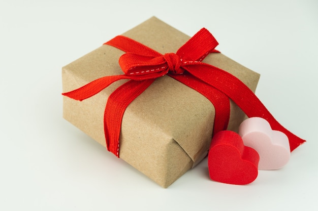 Primer plano de una caja de regalo con cinta roja y dos corazones sobre fondo blanco.