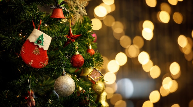 Primer plano del calcetín rojo y blanco de la decoración de la víspera de Navidad que cuelga decorando en la rama de un árbol de pino de Navidad verde con una bola de esfera de palo brillante y una campana en el frente borrosa de fondo dorado con poca luz bokeh.