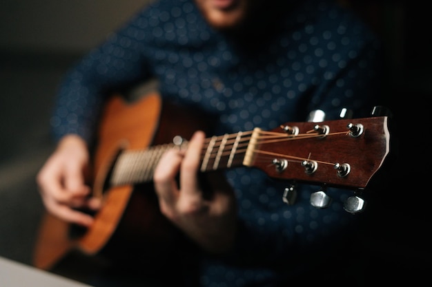 El primer plano de un cantante guitarrista irreconocible tocando la guitarra acústica sentado en un sillón en un salón oscuro con enfoque selectivo