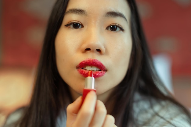 El primer plano en la cara de una hermosa mujer asiática poniendo lápiz labial rojo en sus labios se enfoca en la boca