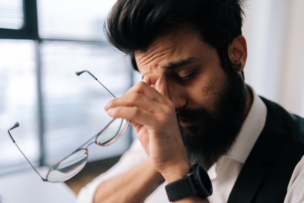 Primer plano de la cara de un hombre de negocios indio cansado y agotado masajeando el puente de la nariz quitándose las gafas sintiendo fatiga visual sentado en el escritorio