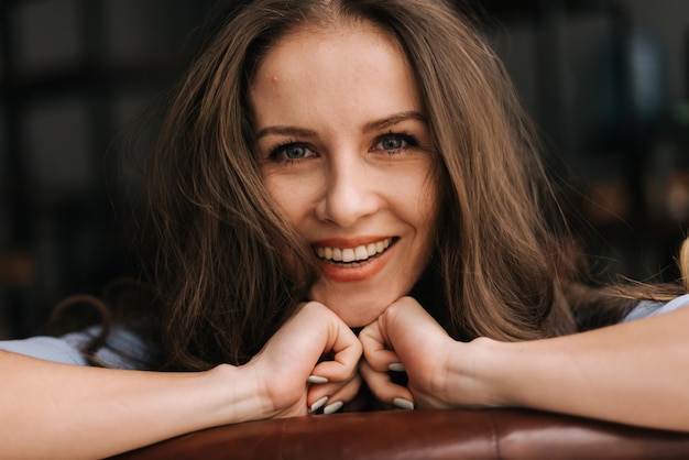 Primer plano de la cara de una joven sonriente sexy con el pelo largo y despeinado sentada y apoyada en el respaldo de la silla. Señora encantadora del negocio que mira la cámara, primer.