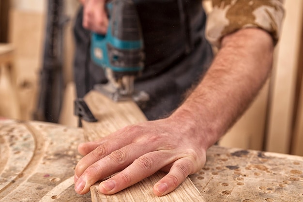 Primer plano de carpintero experimentado en ropa de trabajo y propietario de una pequeña empresa carpintero vio y procesa los bordes de una barra de madera con una sierra caladora en un taller ligero