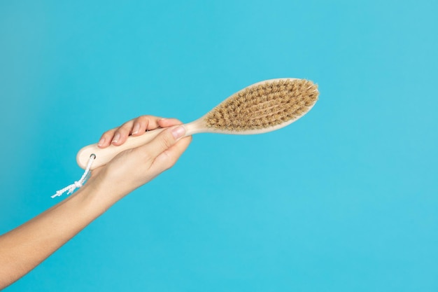 Primer plano de cepillo de masaje de piel seca hecho de madera en mano femenina sobre fondo azul Procedimientos anticelulíticos de autocuidado y cuidado corporal destinados a deshacerse de la celulitis