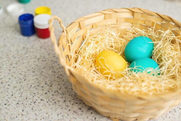 Foto primer plano de una cesta de mimbre en la que hay huevos pintados hay pinturas en la mesa cercana