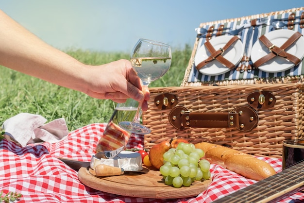 Primer plano de una cesta de picnic con bebidas y comida en la hierba mano masculina sosteniendo una copa de vino