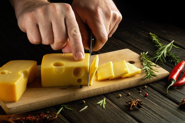 Primer plano de un chef con las manos con un cuchillo cortando queso en una tabla de cortar en la cocina de un restaurante cocinando productos lácteos para el almuerzo durante una dieta Espacio negro para receta