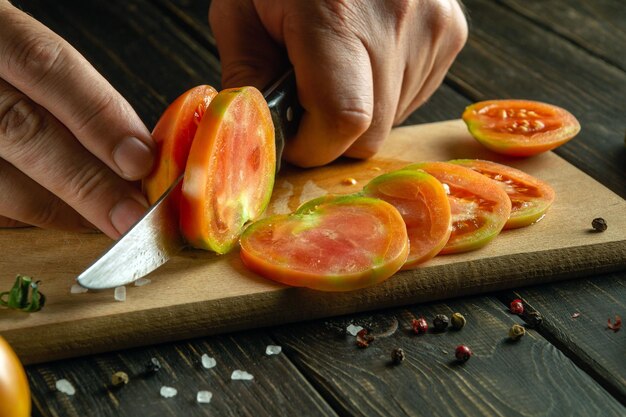 Primer plano de un chef con las manos con un cuchillo cortando un tomate en pedazos pequeños para una ensalada de verduras Ambiente de trabajo en la mesa de la cocina