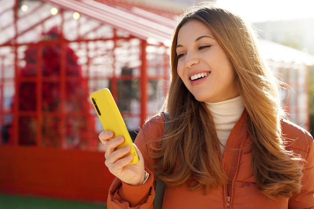 Primer plano de una chica alegre con chaqueta acolchada usando un teléfono inteligente al aire libre