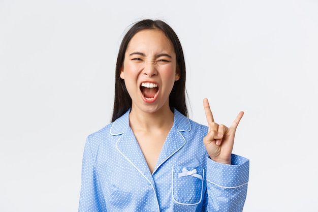 Primer plano de una chica asiática atractiva rebelde y despreocupada en pijama azul que tiene una fiesta de pijamas, que muestra un gesto de rock-n-roll como enloquecido, gritando sí y disfrutando de la música, fondo blanco.