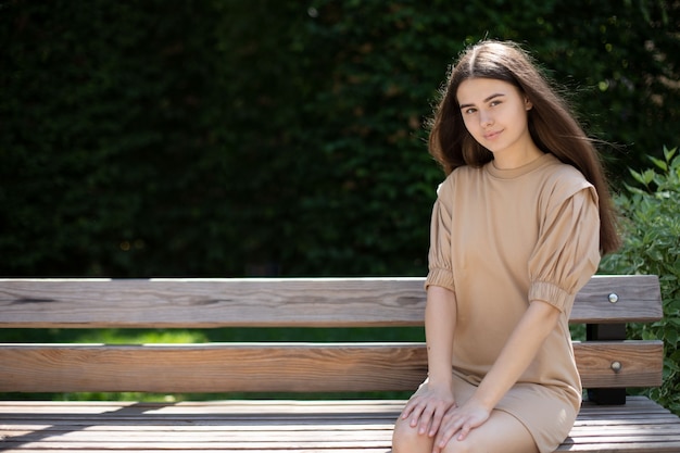 Primer plano de una chica morena con un vestido beige con el pelo largo se sienta en un banco de madera en el parque con un fondo de vegetación. lugar para tu diseño