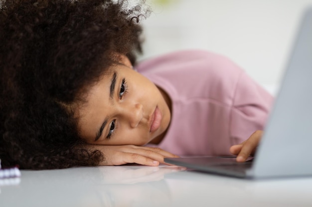 El primer plano de una chica negra aburrida acostada en el escritorio con una laptop