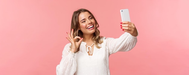 El primer plano de una chica rubia guapa y satisfecha con vestido blanco tomando una selfie graba un video del teléfono móvil muestra un signo satisfactorio con un asentimiento complacido sonriendo de acuerdo o recomienda un fondo rosa