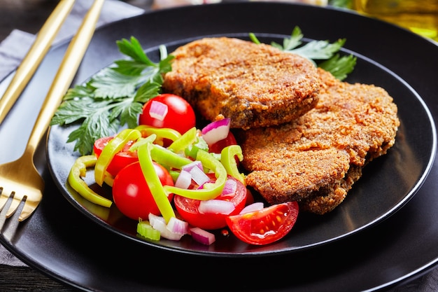 Primer plano de chuletas de cordero empanizadas servidas con tomate, cebolla roja, ensalada de pimiento verde en un plato negro sobre una mesa de madera con tenedor y cuchillo dorado, vista horizontal