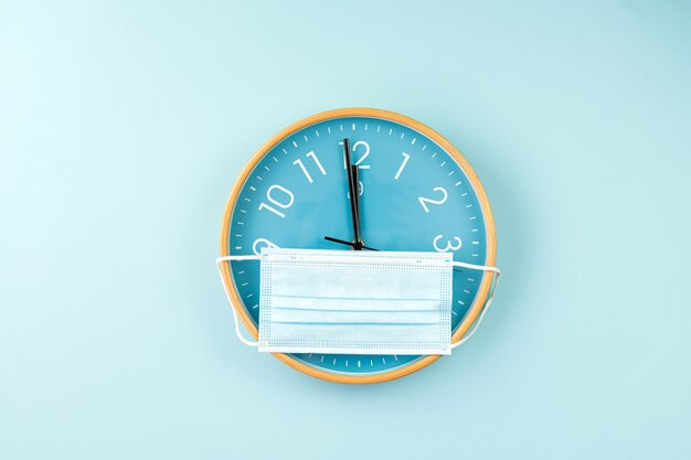 Primer plano de un colorido reloj de pared sobre fondo azul. Imagen minimalista plana de reloj de pared de plástico con máscara médica.