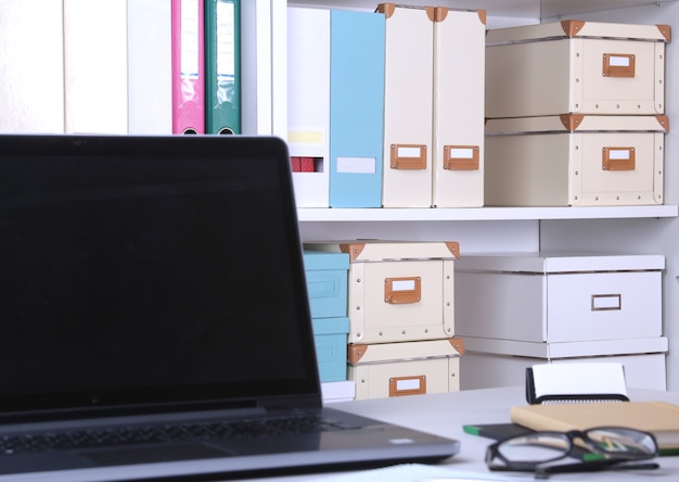 Primer plano de un cómodo lugar de trabajo en la oficina con ordenador portátil, ratón, cuaderno, gafas, bolígrafo y otros equipos en la mesa sobre fondo borroso de muebles.