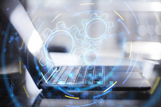 Primer plano de la computadora portátil con holograma de mecanismo de engranaje de hud azul brillante creativo abstracto sobre fondo borroso Concepto de tecnología y trabajo en equipo industrial Doble exposición