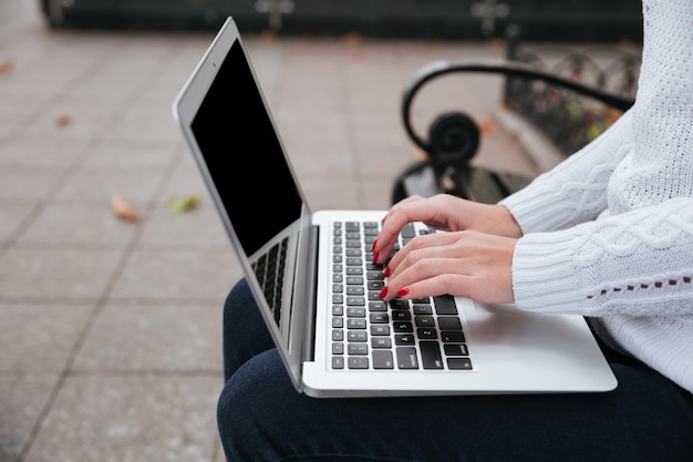 Primer plano de una computadora portátil de pantalla en blanco utilizada por manos de mujer joven sentada en un banco al aire libre