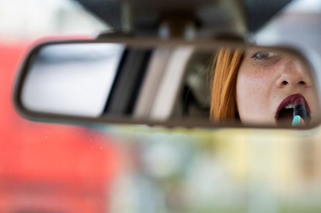 Primer plano de un conductor de mujer joven pelirroja corrigiendo su maquillaje con lápiz labial rojo oscuro mirando en el espejo retrovisor del coche detrás del volante de un vehículo.