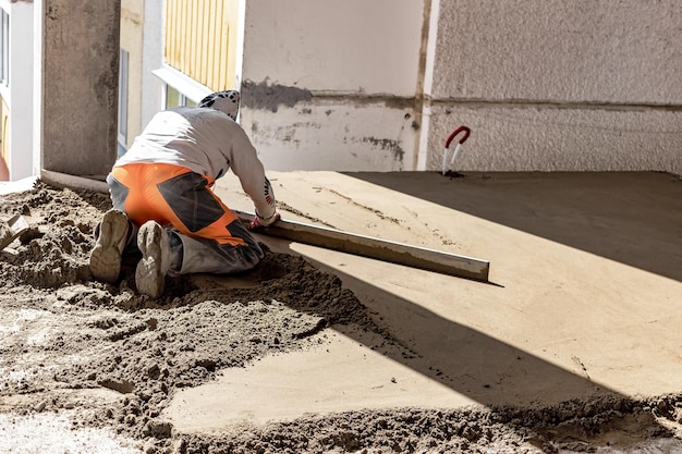 Primer plano del constructor de hombres colocando el riel de la regla en el piso cubierto con una mezcla de arena y cemento en el sitio de construcción. Trabajador de sexo masculino que nivela la superficie con un borde recto mientras nivela el piso. fondo borroso.