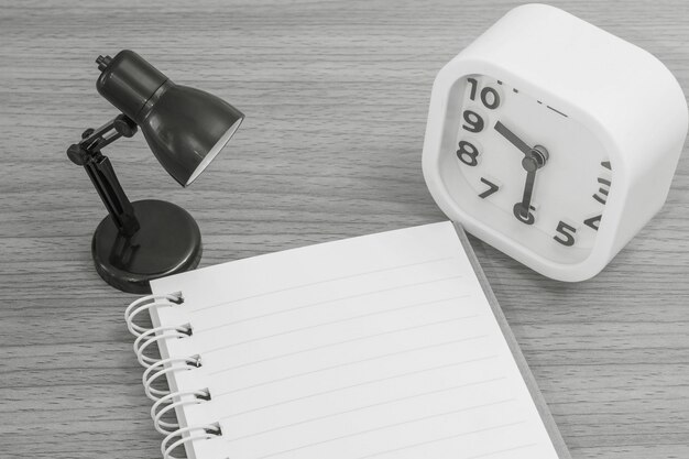 Primer plano de un cuaderno con una pequeña lámpara y un reloj de alarma blanco en el escritorio de madera con textura de fondo en tono blanco y negro