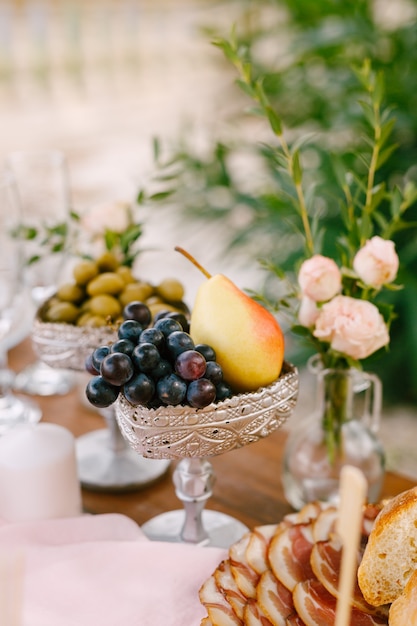 Un primer plano de un cuenco de color plateado con uvas y una pera sobre la mesa con jamón un jarrón con una flor