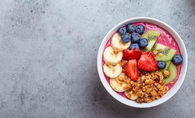 Primer plano de cuencos de batido de acai de verano con fresas, plátano, arándanos, kiwi y granola sobre fondo de hormigón gris. Tazón de desayuno con frutas y cereales, vista superior, espacio para texto