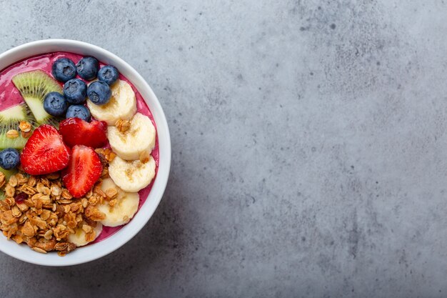 Primer plano de cuencos de batido de acai de verano con fresas, plátano, arándanos, kiwi y granola sobre fondo de hormigón gris. Tazón de desayuno con frutas y cereales, vista superior, espacio para texto