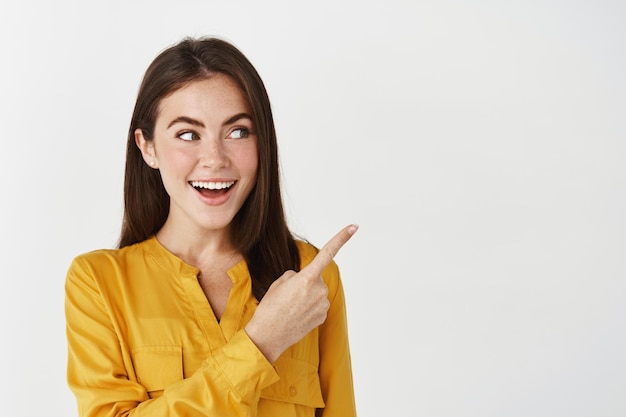 Primer plano de una dama sonriendo, señalando y mirando a la derecha con cara de sorpresa, de pie sobre una pared blanca.