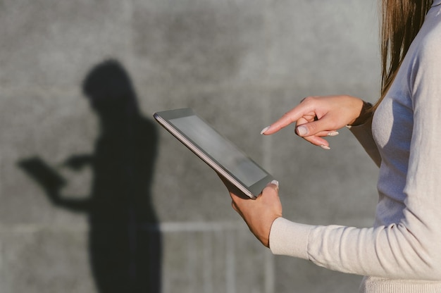 Foto primer plano, un dedo frente a la pantalla de una tableta, en manos de una mujer de negocios. sobre el fondo de un muro de hormigón gris, queda una silueta con una clara sombra.