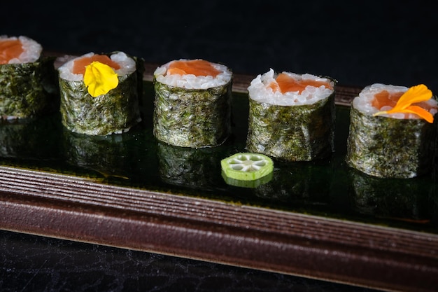 Primer plano de deliciosos rollos de sushi con salmón servidos en bandeja con flores colocadas en una mesa de madera en un restaurante contra un fondo negro