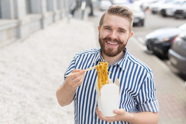 Primer plano de un divertido chico joven inconformista comiendo fideos chinos con palillos de madera sentado en un parque al aire libre en un cálido día de verano. El concepto de descanso y picoteo en la calle.