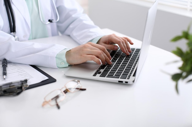 Primer plano de una doctora escribiendo en una computadora portátil, sentada en la mesa del hospital.