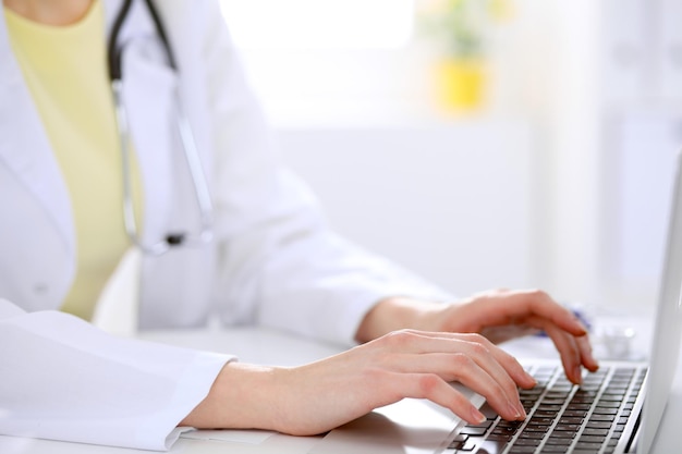 Primer plano de una doctora escribiendo una laptop sentada en una mesa en el hospital.