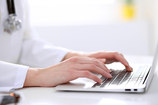 Primer plano de una doctora escribiendo una laptop sentada en una mesa en el hospital.