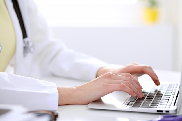 Primer plano de una doctora escribiendo una laptop sentada en una mesa en el hospital.