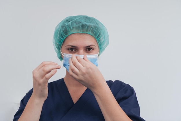 Primer plano de una doctora o científica cansada con una máscara médica y un gorro quirúrgico sobre fondo gris. El auténtico héroe médico real de blanco en la pandemia del coronavirus se está ajustando la máscara.