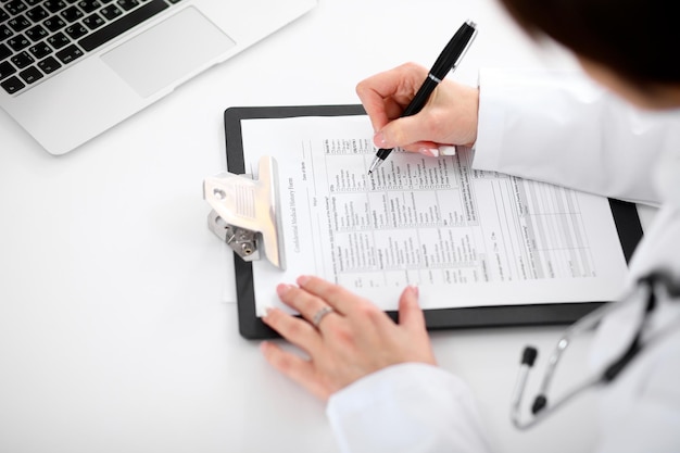 Primer plano de una doctora que llena el formulario de solicitud, sentada en la mesa del hospital