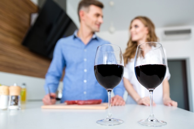 Foto primer plano de dos copas de vino tinto y la feliz pareja de pie en la cocina