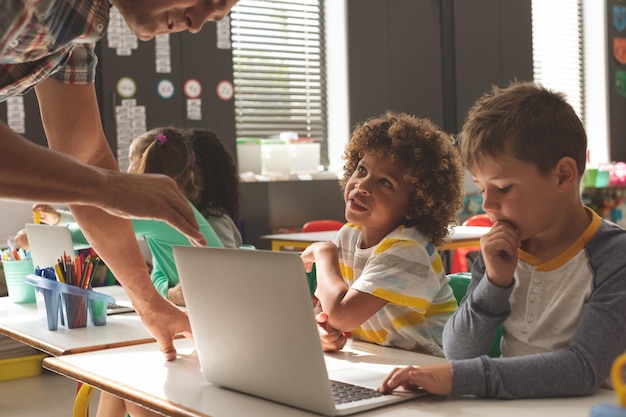 Primer plano de dos escolares escuchando a un maestro explicándoles cómo usar una computadora portátil