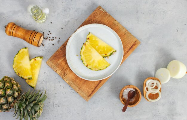 Primer plano de dos piezas de piñas amarillas en rodajas en un plato blanco en una tabla de cortar de madera colocada en la mesa con otros ingredientes aceite aromático en un vial de vidrio botella de pimienta sal en un tazón de madera y cebolla