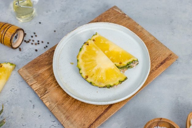 Primer plano de dos piezas de piñas amarillas en rodajas en un plato blanco en una tabla de cortar de madera colocada en la mesa con otros ingredientes aceite aromático en un vial de vidrio botella de pimienta sal en un tazón de madera y cebolla