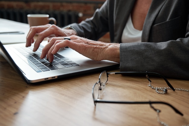 Primer plano de la empresaria irreconocible sentada a la mesa con gafas y preparando la presentación en la computadora portátil