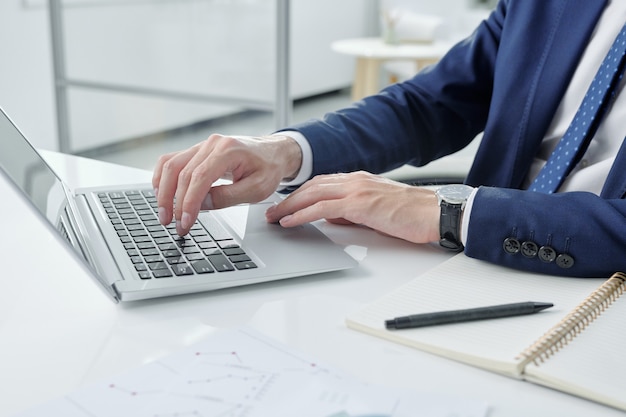 Primer plano del empresario irreconocible en chaqueta sentado en el escritorio con el bloc de notas y escribiendo en la computadora portátil en la oficina