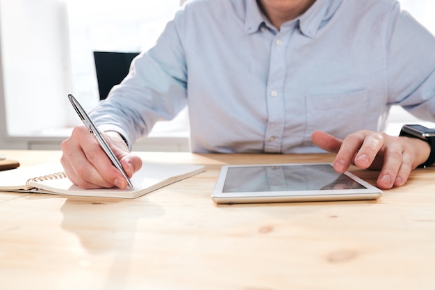 Foto primer plano del empresario irreconocible sentado en el escritorio y usando la tableta mientras toma notas sobre las estadísticas de promoción