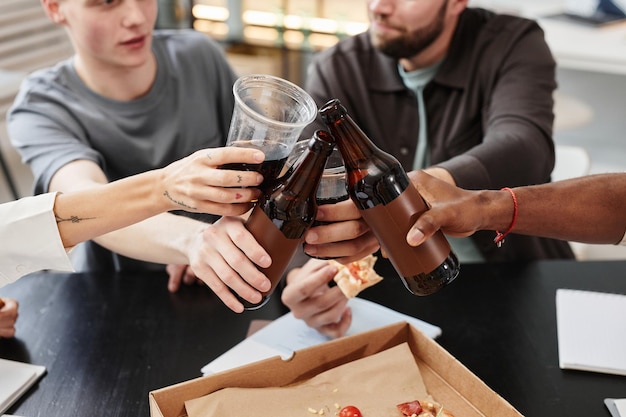 Primer plano de empresarios brindando con botellas de cerveza mientras se sientan en la mesa en la oficina después del trabajo y celebran un día exitoso