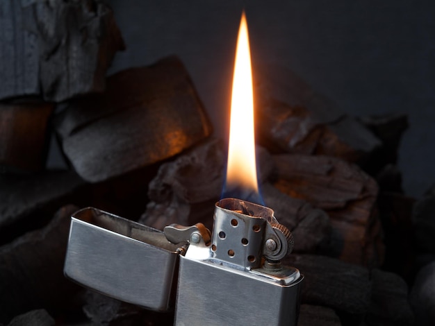 Foto primer plano de un encendedor de gas metálico con fuego ardiente sobre fondo de carbón oscuro con reflejos