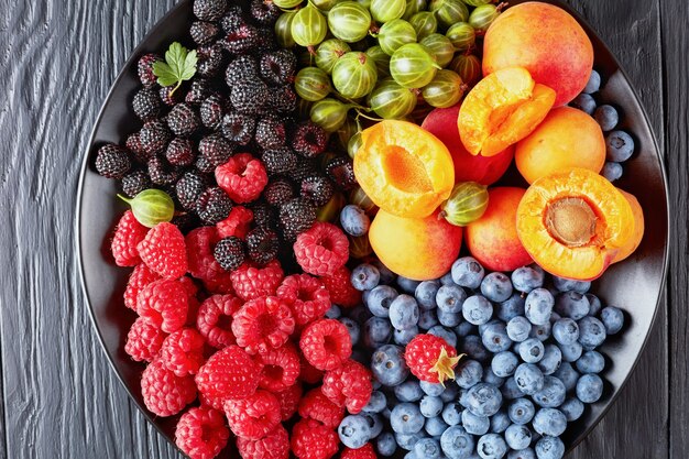 Primer plano de ensalada de frutas y bayas de grosella orgánica fresca, frambuesa roja y negra, arándano, rodajas de albaricoque en una placa negra sobre una mesa de madera, vista horizontal desde arriba, endecha plana